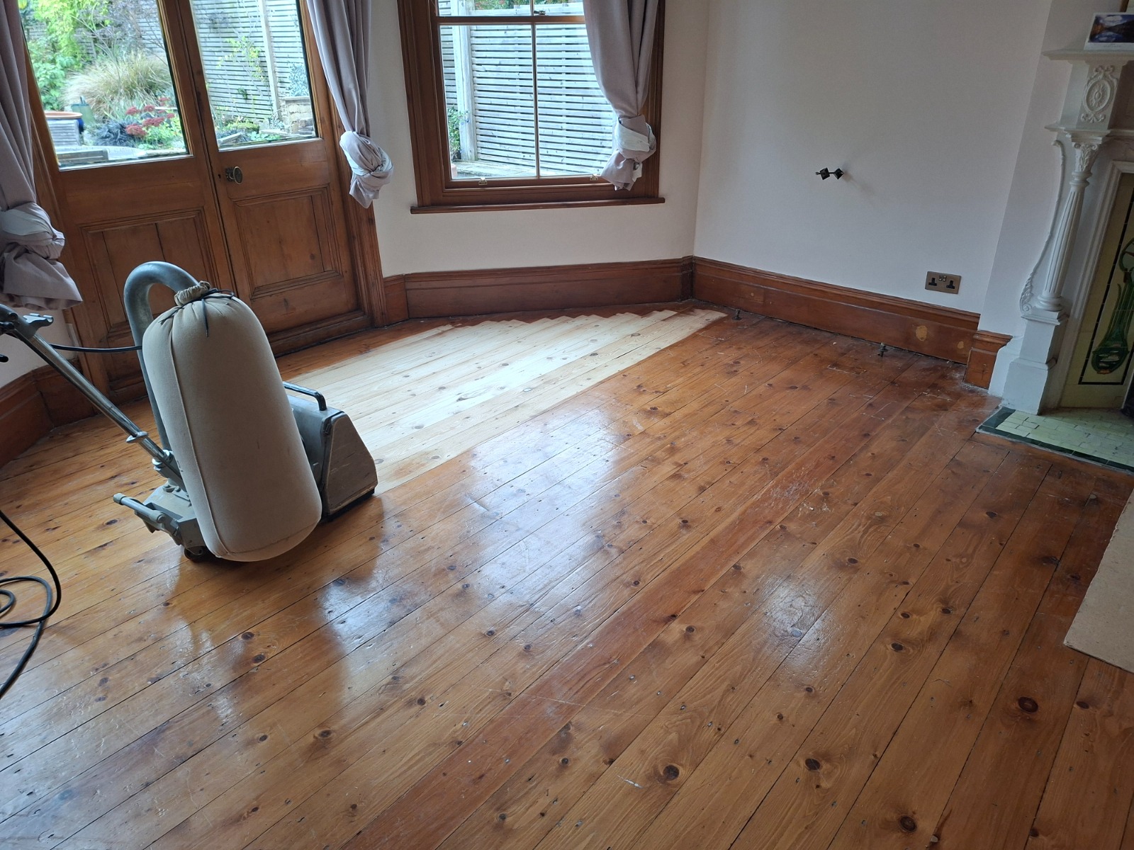 Clapham Floor Sanding in progress on Victorian pine softwood floorboards in a Clapham living room, showing a drum sander and a clear contrast between the freshly sanded pale boards and the old, dark finish.