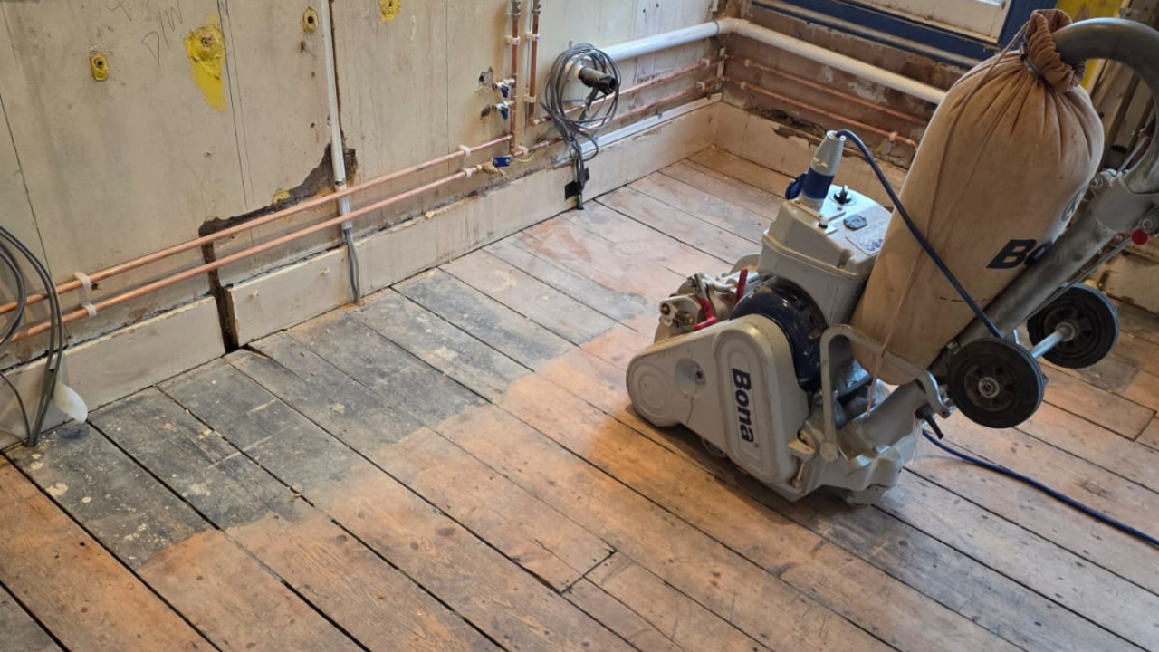 Wood Floor Sanding in Clapham kitchen showing original pine softwood floorboards being sanded with a professional dust-extracting machine, with part of the timber already cleaned back to bare wood.